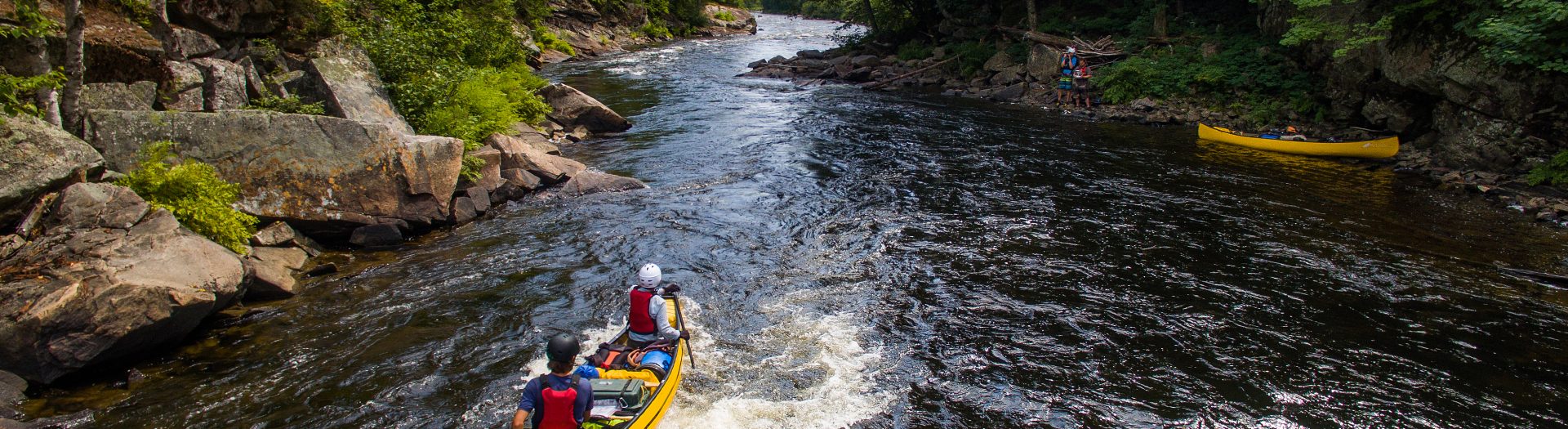 Two yellow canoes being paddles down river through rapids. Trees and rocks on shore.
