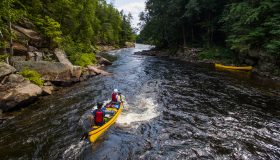 Two yellow canoes being paddles down river through rapids. Trees and rocks on shore.