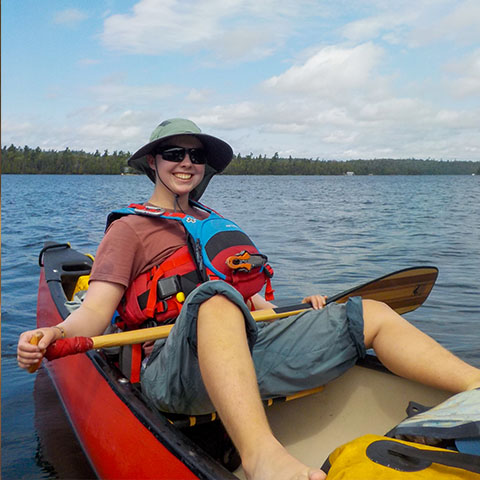 Image of aperson in a hat and lifejacket, sitting in the back of a canoe