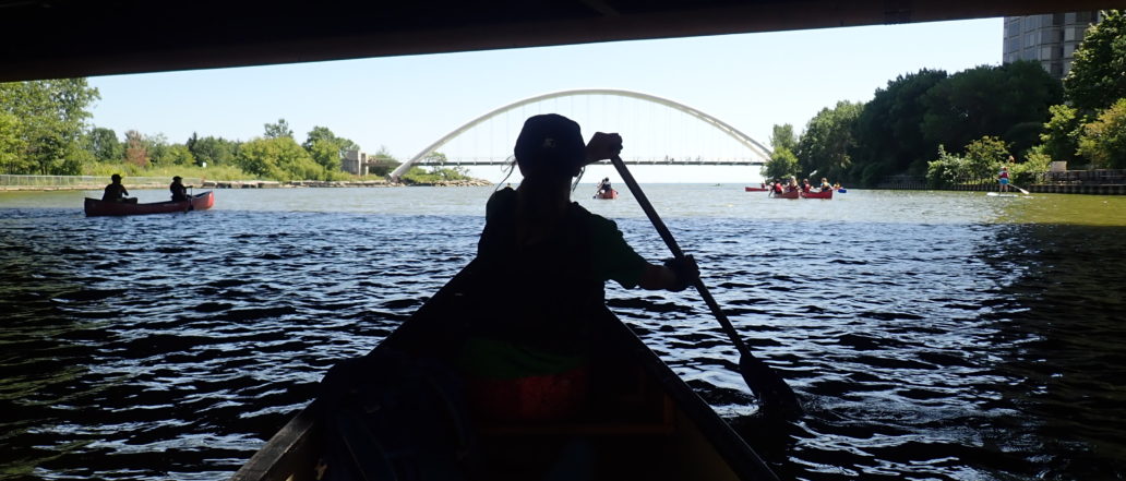 Silouette of a person canoeing under a bridge with a suspension bridge in front of them