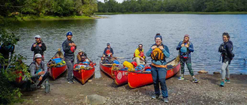 Image of a group of instructors at Ontario staff training. The group is on the shore of the lake with a handful of canoes.