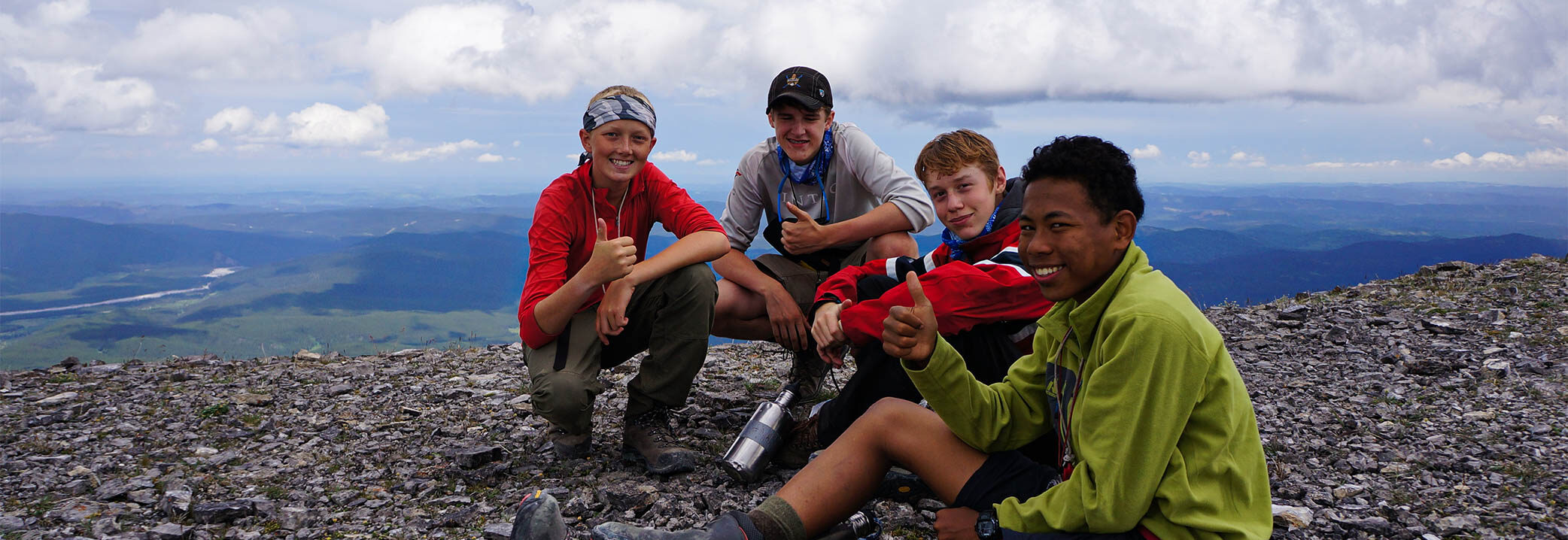 Image of four teens sitting on the top of mountains showing thumbs up