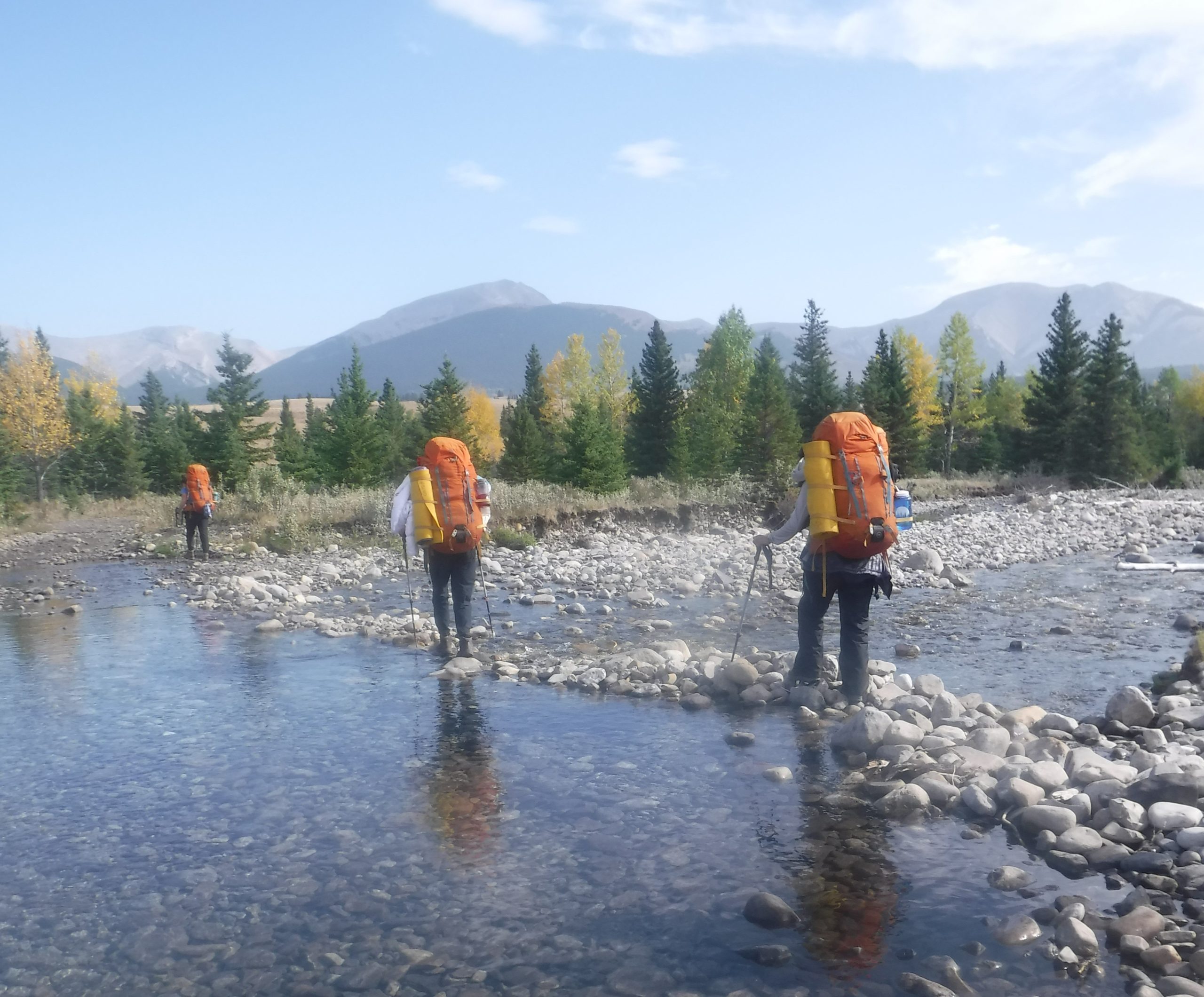 Three people crossing rocky stream with backpacks, mountains and trees in the distance