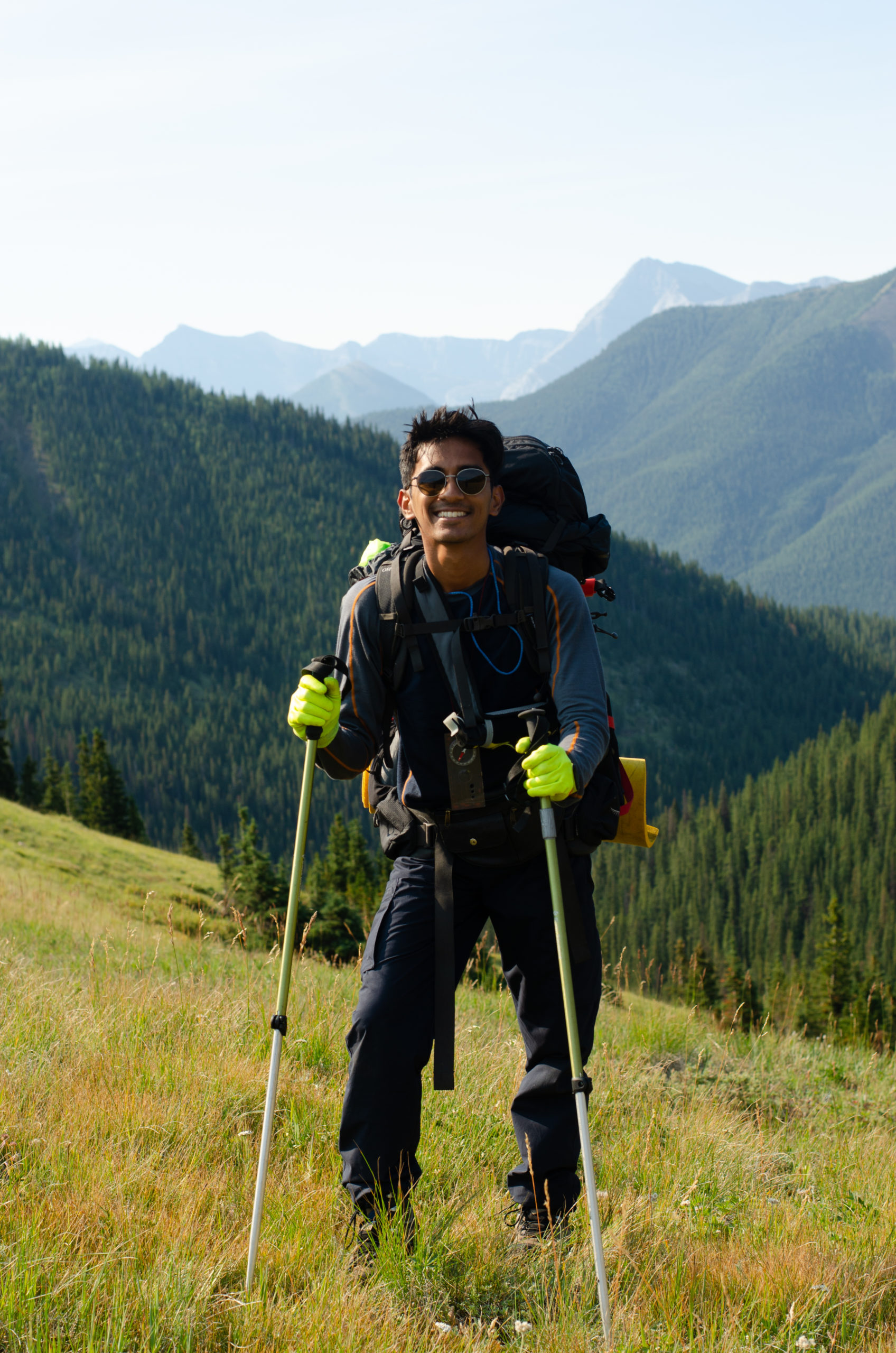 Image of Aryan standing in front of tree covered mountain. He is holding 2 hiking poles.